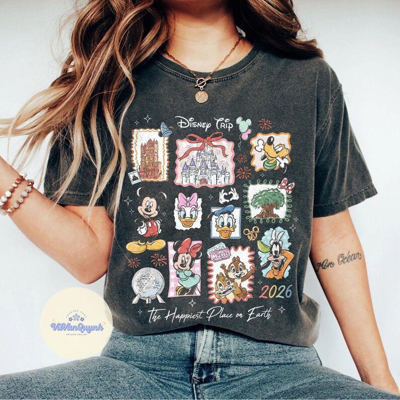 K&ouml;nnte beinhalten: Dunkelgraues T-Shirt mit einem farbenfrohen Disney Trip-Design. Das Design zeigt verschiedene Disney-Charaktere und Wahrzeichen, mit dem Text "The Happiest Place on Earth" und dem Jahr "2026".