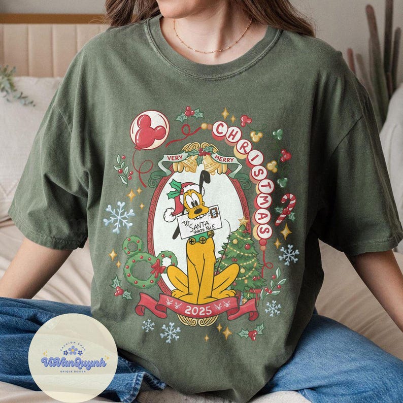 May include: Olive green t-shirt featuring a Christmas design with Pluto holding a sign that reads "To Santa North Pole". The design includes a Christmas tree, snowflakes, and the text "Merry Christmas 2025".
