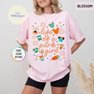 May include: A light pink t-shirt with the phrase "life of a park girl" and various graphics, including a castle, coffee cup, and Mickey Mouse-themed treats. The shirt has a relaxed fit and short sleeves.
