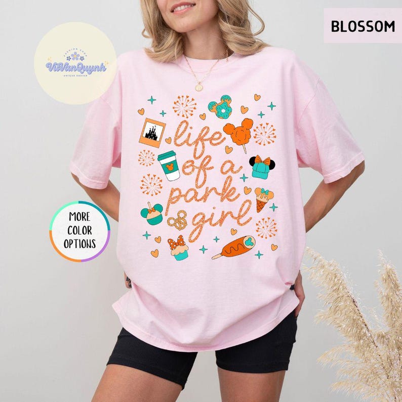 May include: A light pink t-shirt with the phrase "life of a park girl" and various graphics, including a castle, coffee cup, and Mickey Mouse-themed treats. The shirt has a relaxed fit and short sleeves.