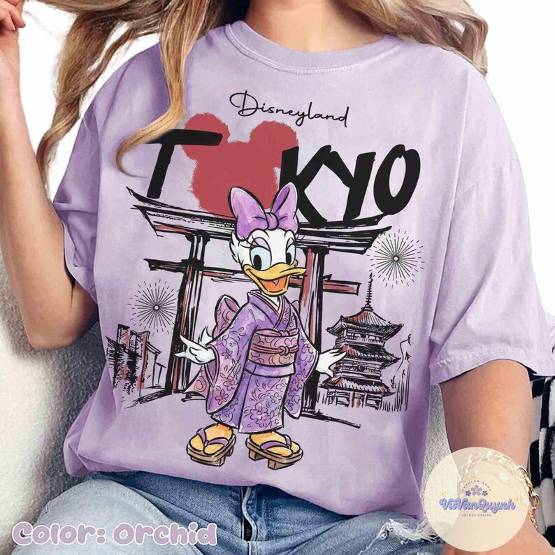 K&ouml;nnte beinhalten: Orchideenfarbenes T-Shirt mit einer Grafik von Daisy Duck in einem Kimono, mit dem Text "Disneyland TOKYO". Das Design umfasst japanische architektonische Elemente und Feuerwerk. Das Shirt hat eine lockere Passform.