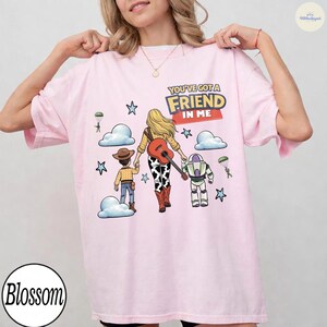 Op de afbeelding: Een lichtroze T-shirt met een afbeelding van een cowboy, een cowgirl met een gitaar en een space ranger. De tekst luidt YOU'VE GOT A FRIEND IN ME. Het ontwerp bevat wolken, sterren en kleine figuren aan parachutes op een lichte achtergrond.