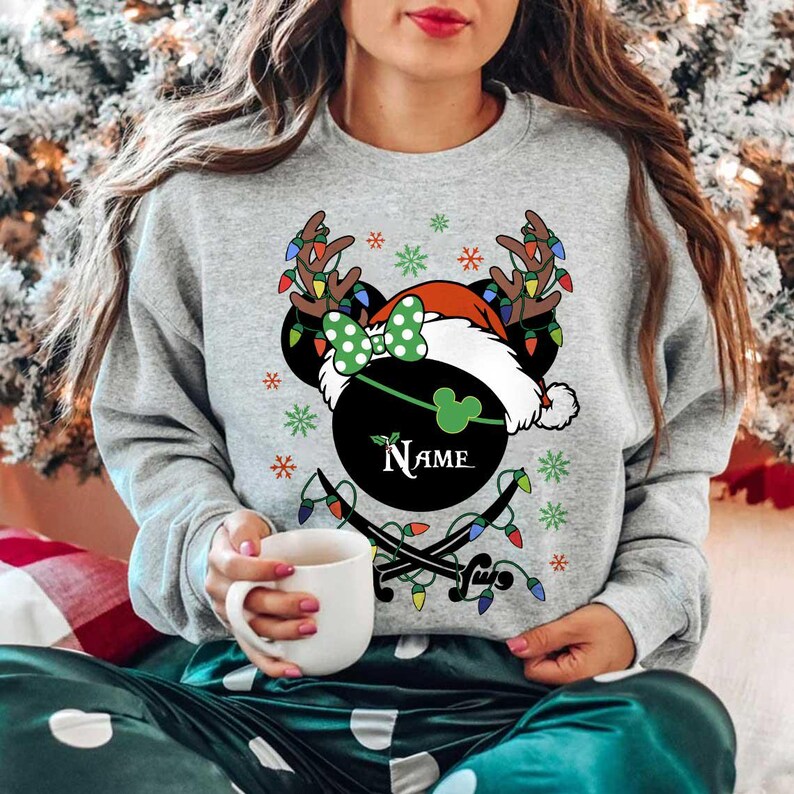May include: A gray sweatshirt featuring a festive design with a black Mickey Mouse head wearing a Santa hat and reindeer antlers adorned with Christmas lights. The design includes a green bow, snowflakes, and the name "Name" in a circular emblem.