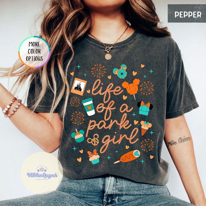 May include: Dark gray t-shirt with the text "life of a park girl" and various cartoon graphics in orange and teal. The shirt also has the words "More Color Options" and "Pepper" on it.