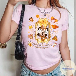 May include: Pink t-shirt featuring a Minnie Mouse graphic in a gold frame with bows and surrounding illustrations. The shirt has a crew neck and short sleeves. The text "THE HAPPINESS IS..." is printed below the frame.