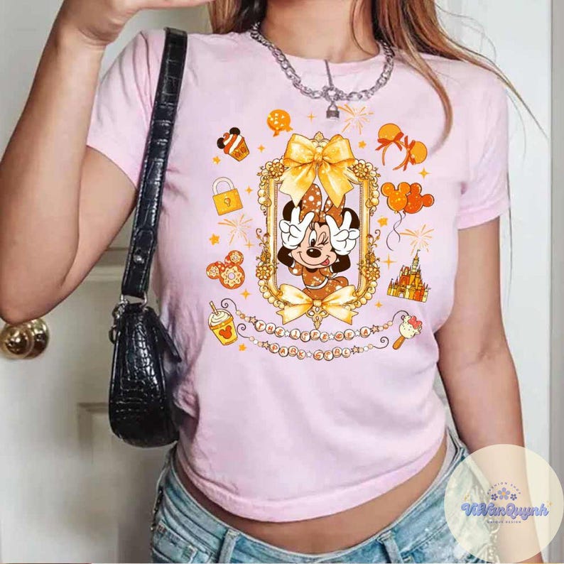 May include: Pink t-shirt featuring a Minnie Mouse graphic in a gold frame with bows and surrounding illustrations. The shirt has a crew neck and short sleeves. The text "THE HAPPINESS IS..." is printed below the frame.