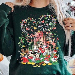 May include: Dark green sweatshirt featuring a festive Disney design. The graphic includes Mickey Mouse ears outlined with Christmas lights, a castle, and several Disney characters in holiday attire. Colorful stars and confetti add to the celebratory theme.