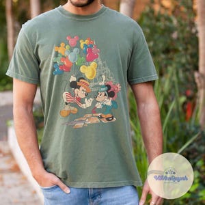 May include: Green t-shirt featuring a colorful graphic of Mickey and Minnie Mouse holding hands, with Mickey Mouse balloons and a castle in the background. The shirt is a muted green color and has a vintage style.