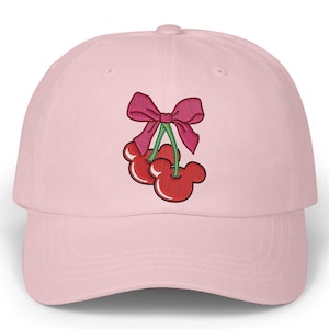 May include: A light pink baseball cap featuring an embroidered design of two red cherries with Mickey Mouse-shaped faces, green stems, and a pink bow. The cap has a curved brim and a rounded crown.