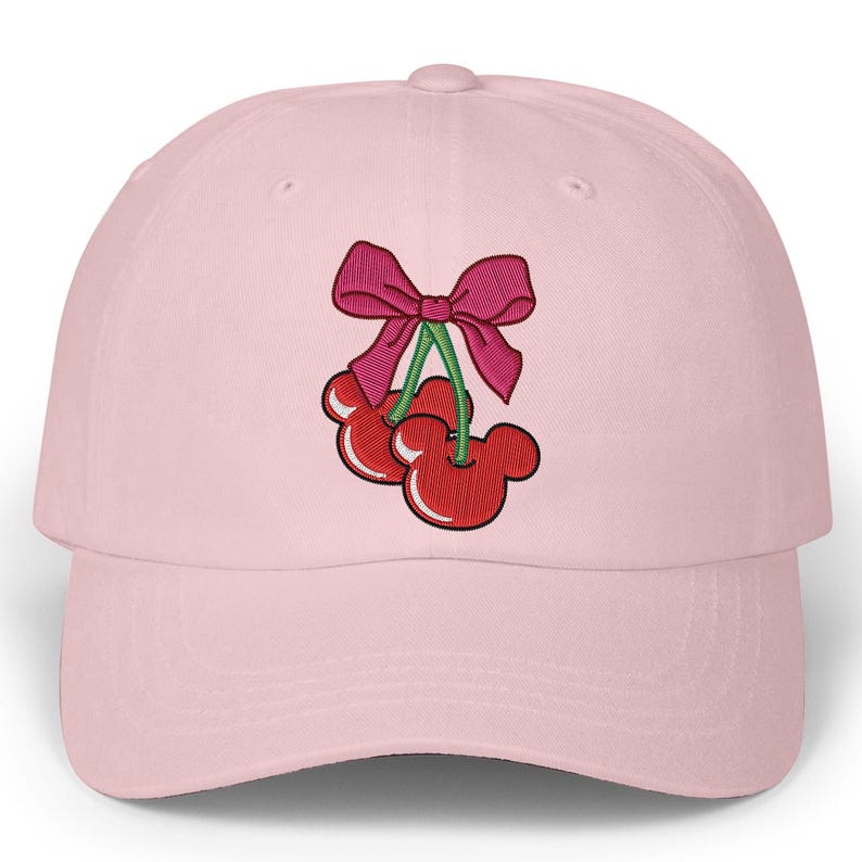 May include: A light pink baseball cap featuring an embroidered design of two red cherries with Mickey Mouse-shaped faces, green stems, and a pink bow. The cap has a curved brim and a rounded crown.
