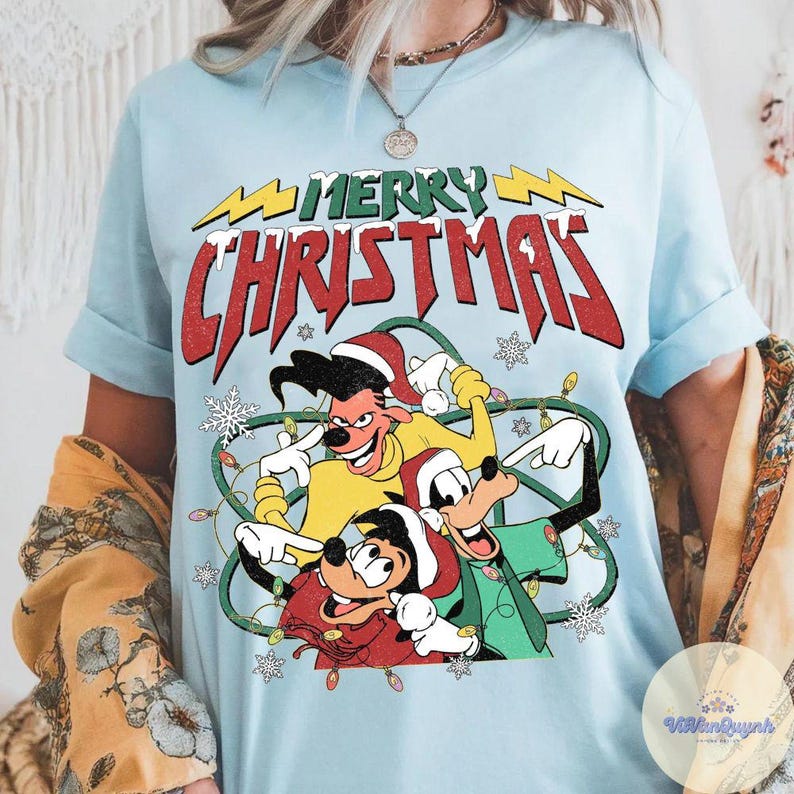 May include: Light blue t-shirt featuring a cartoon graphic with the text "MERRY CHRISTMAS". The graphic includes three cartoon characters wearing Santa hats and surrounded by Christmas lights and snowflakes.