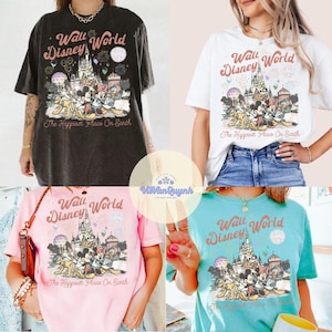 K&ouml;nnte beinhalten: Vier kurz&auml;rmelige T-Shirts in verschiedenen Farben: Schwarz, Wei&szlig;, Rosa und T&uuml;rkis. Jedes Shirt zeigt eine Grafik von Disney-Figuren vor einem Schloss mit dem Text "Walt Disney World" und "The Happiest Place On Earth".
