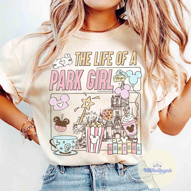 May include: Cream-colored t-shirt with the text "THE LIFE OF A PARK GIRL" in a pink and white design. The graphic includes cartoon images of a castle, popcorn, a teacup, and other park-themed elements.