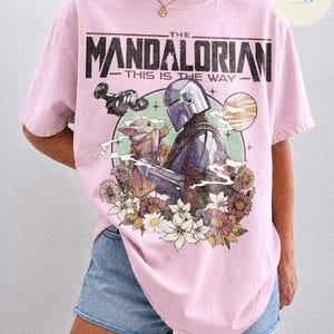 K&ouml;nnte beinhalten: Hellrosa T-Shirt mit einem Aufdruck von The Mandalorian und Grogu, umgeben von Blumen und einem Planeten. Der Text "THE MANDALORIAN - THIS IS THE WAY" ist oben aufgedruckt. Das T-Shirt ist mit "COMFORT COLORS BLOSSOM" gekennzeichnet.