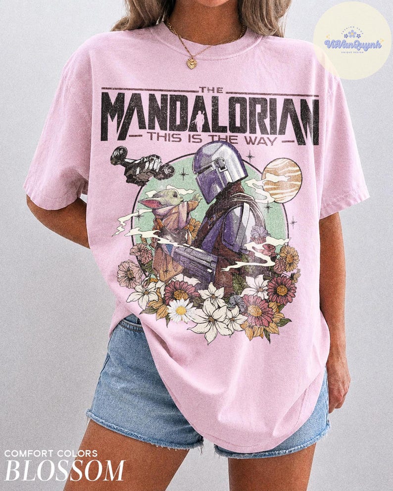 K&ouml;nnte beinhalten: Hellrosa T-Shirt mit einem Aufdruck von The Mandalorian und Grogu, umgeben von Blumen und einem Planeten. Der Text "THE MANDALORIAN - THIS IS THE WAY" ist oben aufgedruckt. Das T-Shirt ist mit "COMFORT COLORS BLOSSOM" gekennzeichnet.