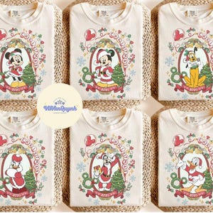 May include: Cream-colored t-shirts with Christmas-themed Disney character graphics. Each shirt features a different character, including Mickey, Minnie, Pluto, Donald, and Goofy, in festive attire, surrounded by Christmas decorations and the year 2025.