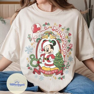 May include: Cream-colored t-shirt featuring a festive Mickey Mouse design. Mickey is dressed in a Santa suit, surrounded by Christmas decorations, the words "VERY MERRY CHRISTMAS" and the year "2025". A holiday-themed graphic tee.