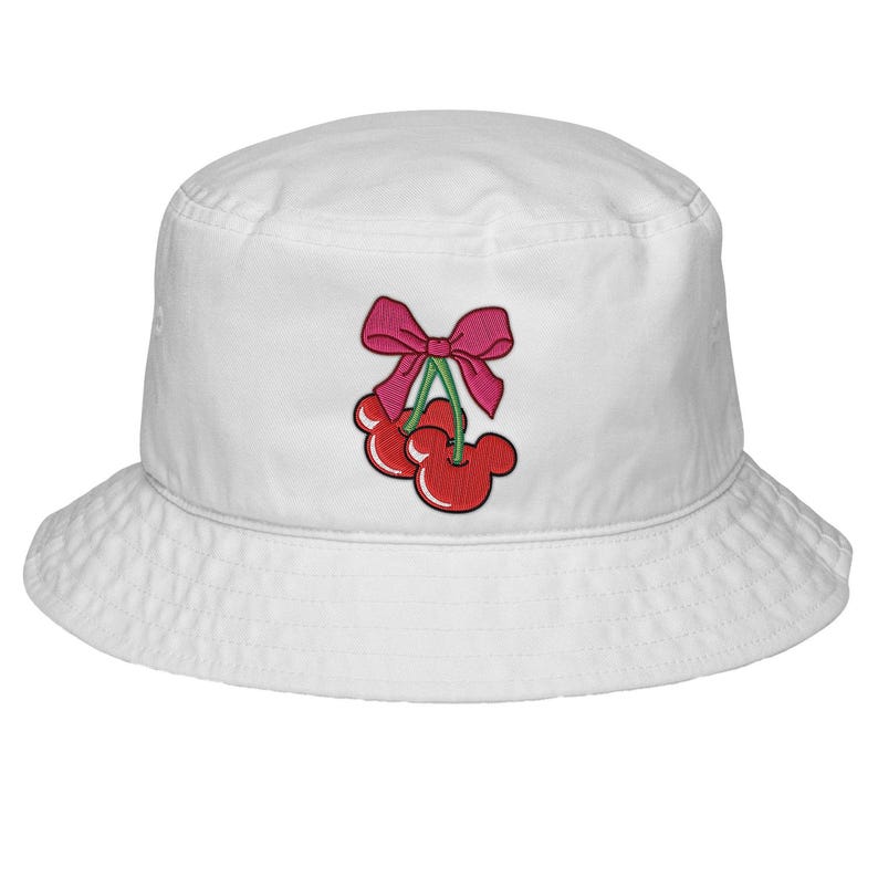 May include: White bucket hat with an embroidered design of red cherries with black outlines and a pink bow. The cherries have a Mickey Mouse ear shape. The hat is made of a soft, durable fabric.