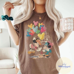May include: Brown t-shirt featuring a vintage-style graphic of Mickey and Minnie Mouse. Mickey Mouse is wearing a straw hat and red and white striped shirt. Minnie Mouse is wearing a blue outfit. Colorful Mickey Mouse balloon graphics are above them.