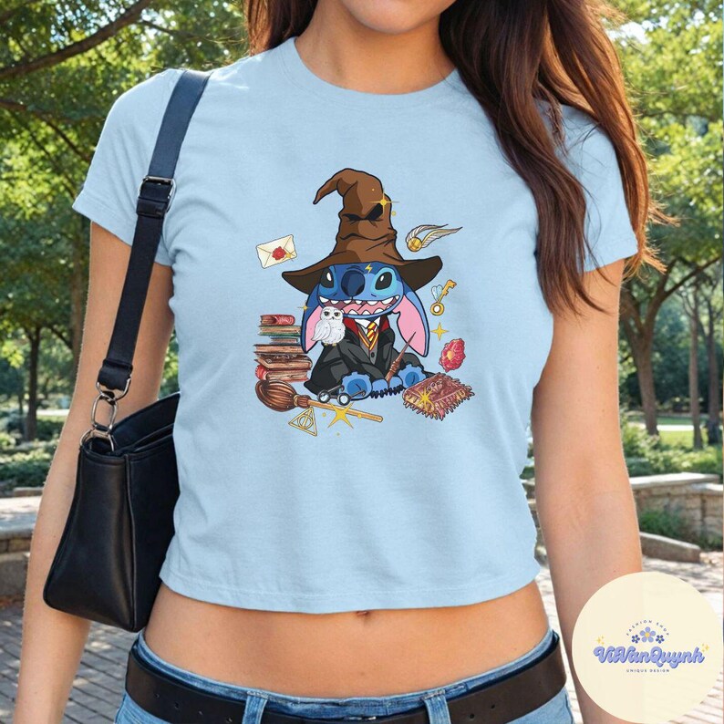 May include: A light blue cropped t-shirt featuring a graphic of Stitch dressed as a wizard. Stitch is wearing a pointed hat, a robe, and holding a wand. He is surrounded by books, a magic spell, and an owl.