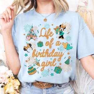 Pu&ograve; includere: T-shirt azzurra con un design grafico colorato. Il design include un castello, Minnie Mouse, regali e la scritta "Life of a birthday girl" in arancione. La maglietta ha scollo tondo e maniche corte.