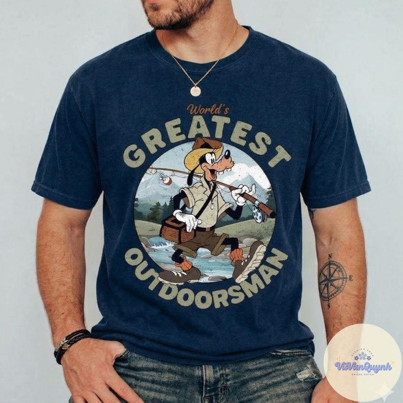 Pu&ograve; includere: T-shirt blu navy con un'illustrazione a cartoni animati di Pippo come pescatore, con la scritta "World's Greatest Outdoorsman" in un design circolare. La grafica include una canna da pesca, una scatola per esche e uno sfondo panoramico.