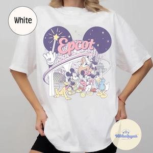 May include: White t-shirt featuring a colorful graphic of Disney characters, including Mickey Mouse, Goofy, and Donald Duck, with the word "Epcot" in pink. The design includes a stylized depiction of a theme park.