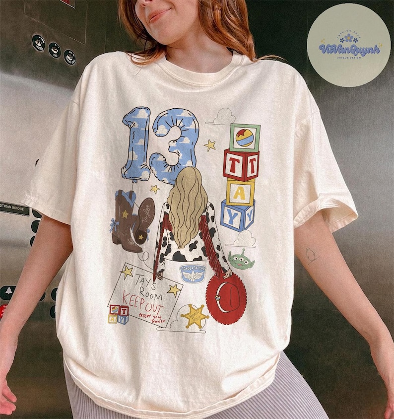 May include: A cream-colored t-shirt featuring a graphic print of the number 13, a person with blonde hair, cowboy boots, alphabet blocks, a green alien, and a sign that reads "Tay's Room KEEP OUT except you Travis". The design includes stars and clouds.