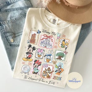 K&ouml;nnte beinhalten: Cremefarbenes T-Shirt mit einem farbenfrohen Disney Trip-Design mit Micky Maus, Minnie Maus, Donald Duck und anderen Charakteren. Das Shirt enth&auml;lt den Text "The Happiest Place on Earth" und das Jahr 2026.