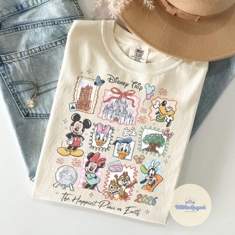 K&ouml;nnte beinhalten: Cremefarbenes T-Shirt mit einem farbenfrohen Disney Trip-Design mit Micky Maus, Minnie Maus, Donald Duck und anderen Charakteren. Das Shirt enth&auml;lt den Text "The Happiest Place on Earth" und das Jahr 2026.