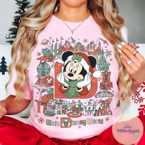 May include: Pink t-shirt featuring a festive Minnie Mouse design with a Santa hat and scarf, surrounded by iconic Walt Disney World landmarks. The text "Walt Disney World" is printed at the bottom in a stylized font.
