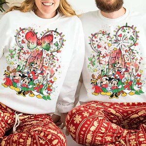 May include: White sweatshirts featuring a colorful Disney holiday design with Mickey Mouse, Minnie Mouse, and other characters. The design includes a castle, lights, and a bow. The subjects are wearing red and white patterned pajama pants.