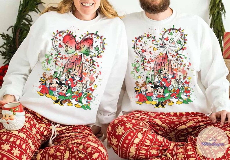 May include: White sweatshirts featuring a colorful Disney holiday design with Mickey Mouse, Minnie Mouse, and other characters. The design includes a castle, lights, and a bow. The subjects are wearing red and white patterned pajama pants.