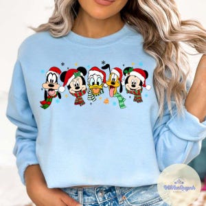 May include: Light blue sweatshirt featuring a festive design with Mickey Mouse, Minnie Mouse, Donald Duck, Goofy, and Pluto. Each character is wearing a Santa hat and a scarf. Snowflakes and a bell are also included in the design.