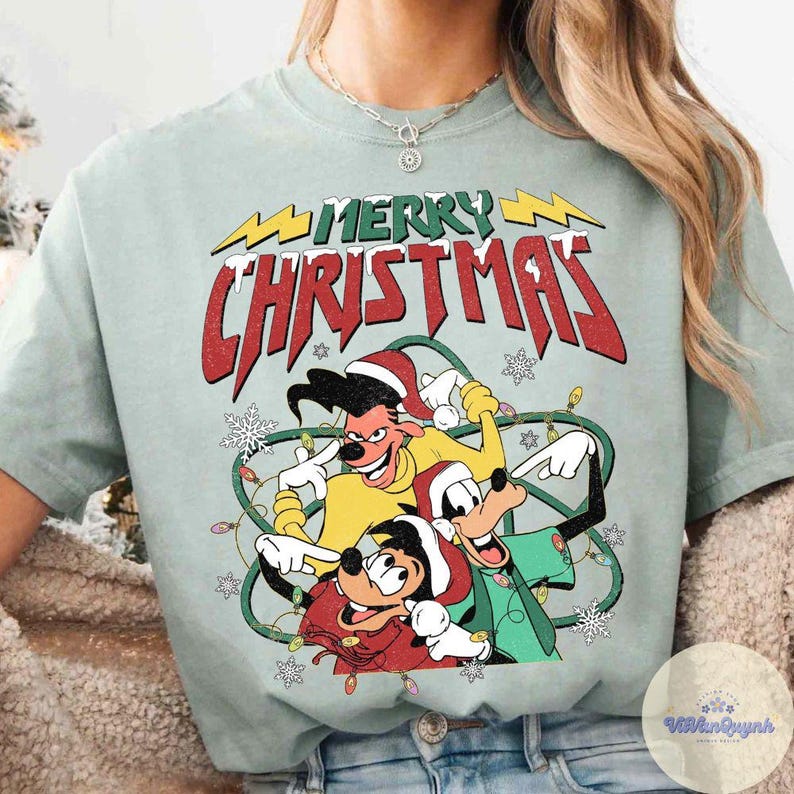 May include: A light green t-shirt featuring a cartoon graphic with the text "MERRY CHRISTMAS". The graphic includes three cartoon characters wearing Santa hats and surrounded by Christmas lights and snowflakes.