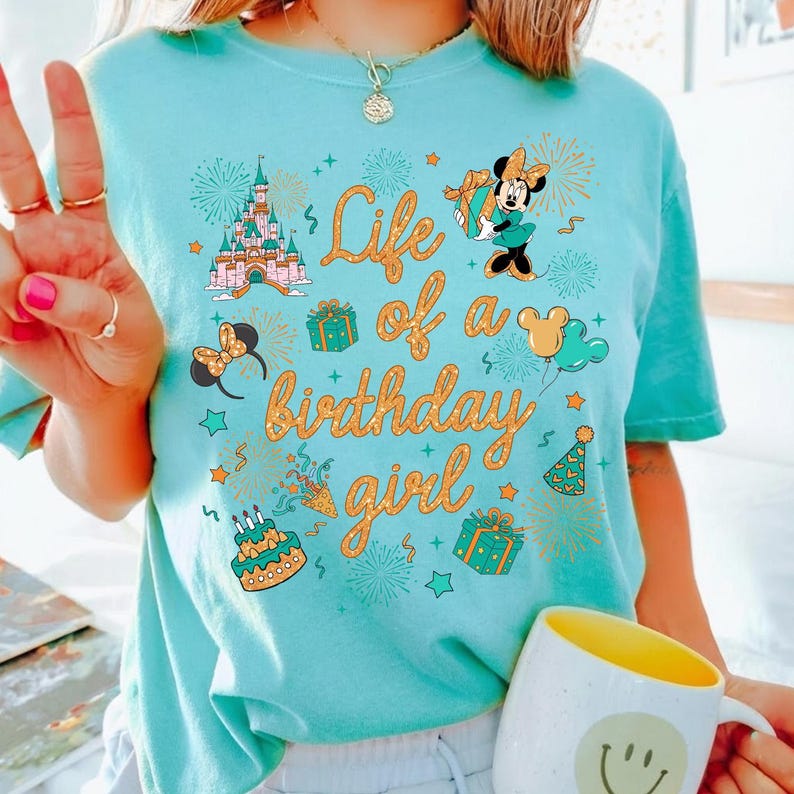 Pu&ograve; includere: Una maglietta azzurra con la scritta dorata "Life of a birthday girl". La maglietta presenta grafiche a cartoni animati di un castello, Minnie Mouse, regali e fuochi d'artificio. &Egrave; visibile una tazza bianca con l'interno giallo.