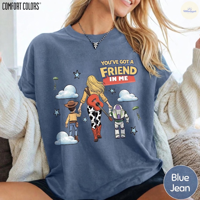 Op de afbeelding: Een blauwgrijs t-shirt met een afbeelding van drie wegwandelende figuren, waaronder een cowboy, een cowgirl met een gitaar en een space ranger. De tekst "YOU'VE GOT A FRIEND IN ME" staat boven de figuren, omringd door wolken en sterren.