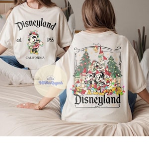 May include: Cream-colored Disneyland t-shirt with front and back graphics. The front features Mickey Mouse in a Santa hat, the text "Disneyland est. 1955 CALIFORNIA". The back shows Mickey and friends in front of a castle and Christmas trees.