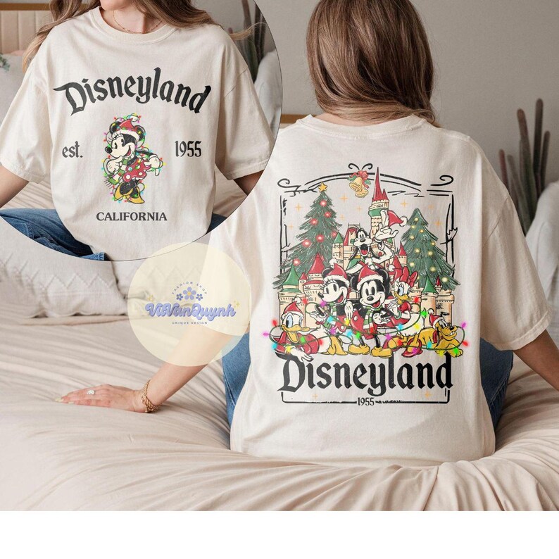 May include: Cream-colored Disneyland t-shirt with front and back graphics. The front features Mickey Mouse in a Santa hat, the text "Disneyland est. 1955 CALIFORNIA". The back shows Mickey and friends in front of a castle and Christmas trees.