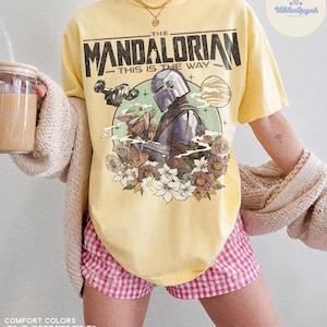 K&ouml;nnte beinhalten: Gelbes T-Shirt mit einem Aufdruck von The Mandalorian und Grogu, mit dem Text "THIS IS THE WAY". Das Design umfasst ein Raumschiff, einen Planeten und Blumen. Das Shirt ist in Buttergelb gehalten.