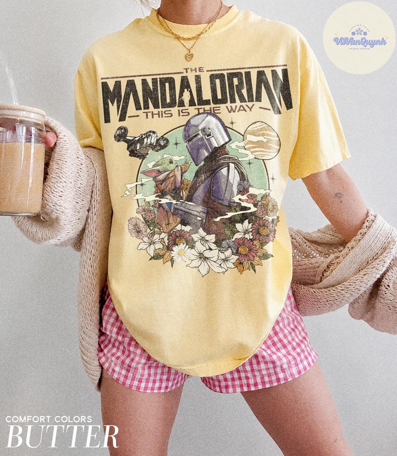 K&ouml;nnte beinhalten: Gelbes T-Shirt mit einem Aufdruck von The Mandalorian und Grogu, mit dem Text "THIS IS THE WAY". Das Design umfasst ein Raumschiff, einen Planeten und Blumen. Das Shirt ist in Buttergelb gehalten.