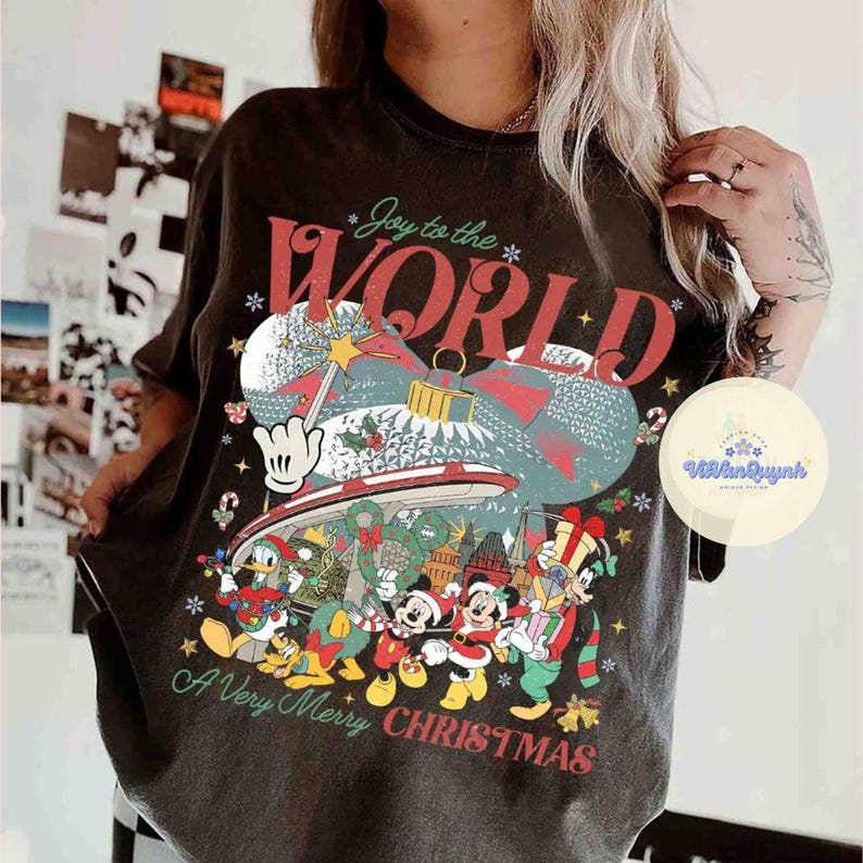 May include: Dark gray t-shirt featuring a festive Christmas design with Mickey Mouse and friends. The shirt has the words "Joy to the World" and "A Very Merry Christmas" in red and white. The graphic includes a Christmas ornament and snowflakes.