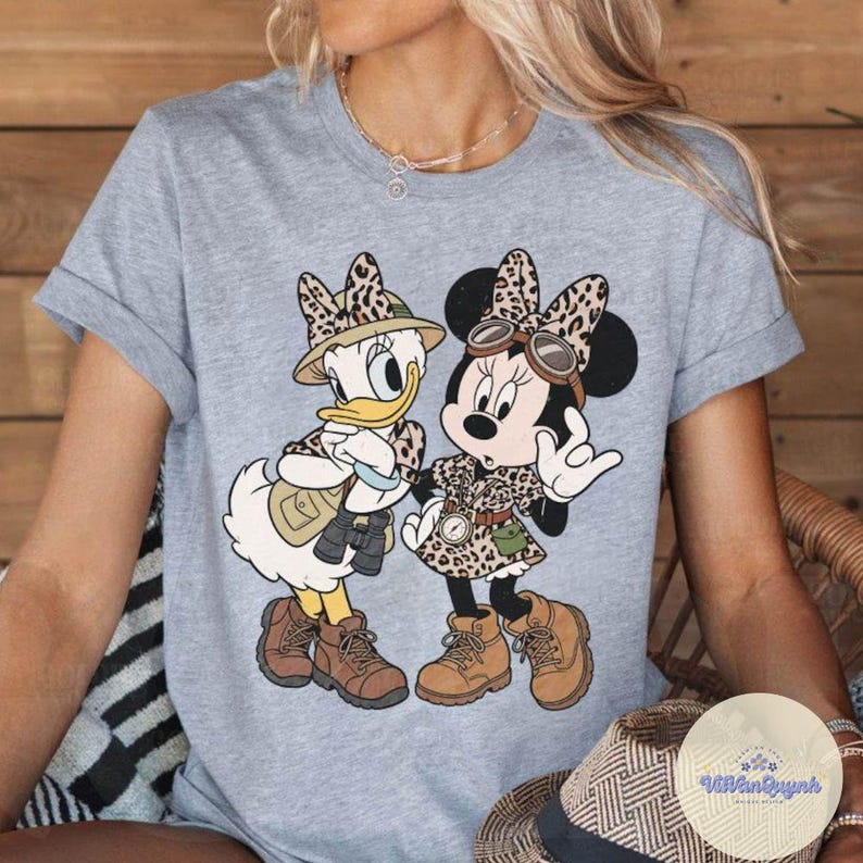 May include: A light gray t-shirt featuring a cartoon illustration of Daisy Duck and Minnie Mouse in safari outfits. Both characters are wearing leopard print clothing, hats, and boots. Daisy Duck is holding binoculars. The shirt has rolled-up sleeves.
