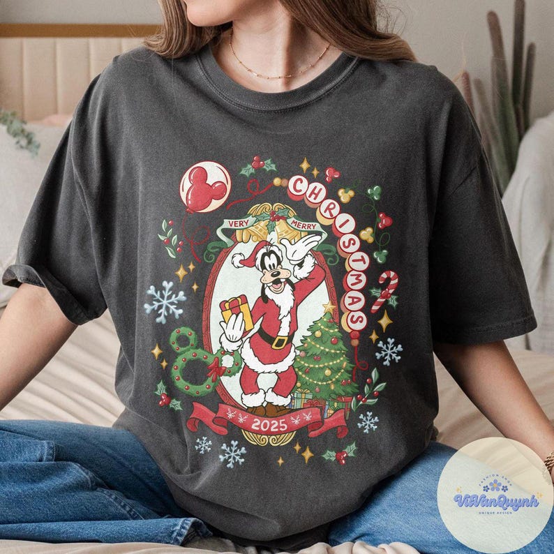 May include: Dark gray t-shirt featuring a Christmas-themed design with the text "Very Merry Christmas 2025." The graphic includes a cartoon character in a Santa suit, a Christmas tree, and festive decorations.