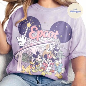 May include: A light purple t-shirt featuring a graphic of Disney characters, including Mickey and Minnie Mouse, Goofy, and Donald Duck. The word "Epcot" is in pink letters. The design includes a wand, stars, and a futuristic building.