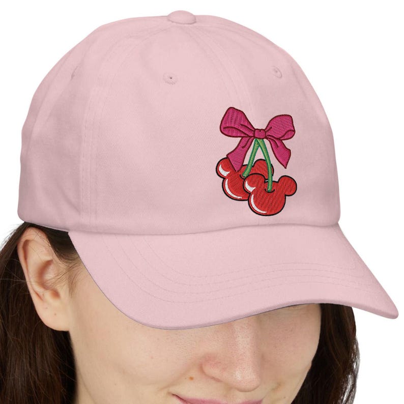 May include: A light pink baseball cap with an embroidered design of two red cherries with Mickey Mouse-shaped outlines, a green stem, and a pink bow. The cap has a curved brim and a rounded crown.