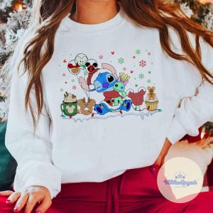 May include: White sweatshirt featuring a colorful cartoon of Stitch in a Santa hat, surrounded by festive holiday characters and snowflakes. The design includes a hot cocoa mug, reindeer, and gingerbread man. A cheerful and whimsical holiday-themed apparel.