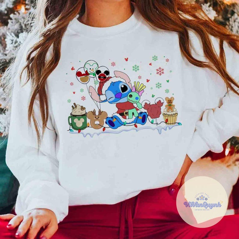 May include: White sweatshirt featuring a colorful cartoon of Stitch in a Santa hat, surrounded by festive holiday characters and snowflakes. The design includes a hot cocoa mug, reindeer, and gingerbread man. A cheerful and whimsical holiday-themed apparel.