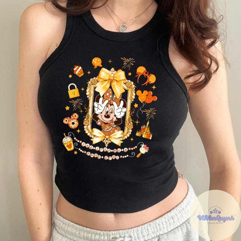 May include: Black tank top featuring a Minnie Mouse graphic in a gold frame. The design includes a bow, fireworks, and other Disney-themed elements. The tank top is a cropped style.