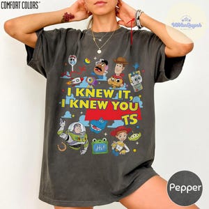 Op de afbeelding: Een antracietgrijs t-shirt met korte mouwen en een kleurrijke grafische print van diverse geanimeerde personages uit de Toy Story-franchise. De tekst luidt I KNEW IT, I KNEW YOU TS in gele en witte letters op een rode achtergrond.